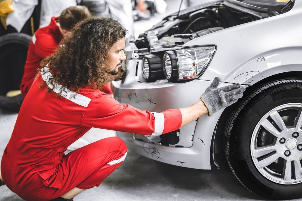 Mechanic in red jumpsuit examining front bumper of a silver car in a garage.