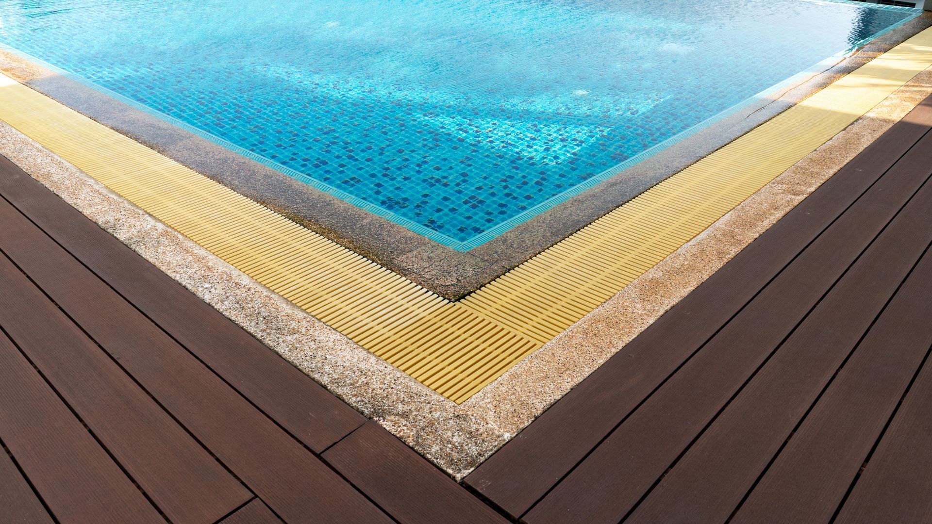 Corner of a turquoise swimming pool with tan stone trim and dark wood deck panels.