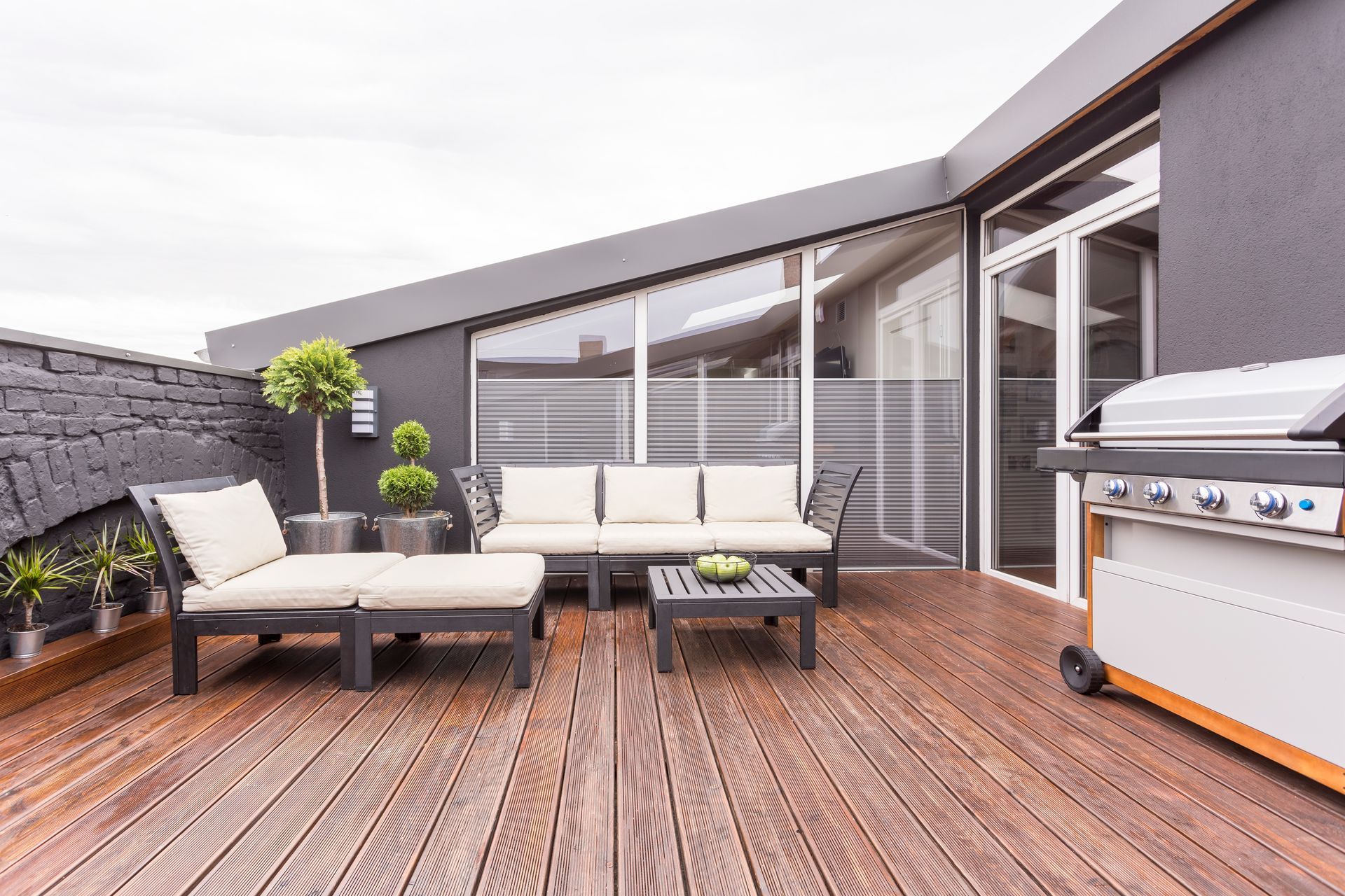 Rooftop terrace with lounge chairs, wooden deck, glass railing, and a gas grill under a bright sky