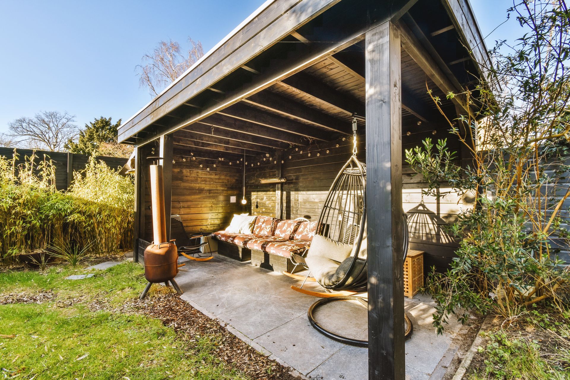 Covered outdoor patio with sofas, string lights, and a wood-fired heater in a garden setting