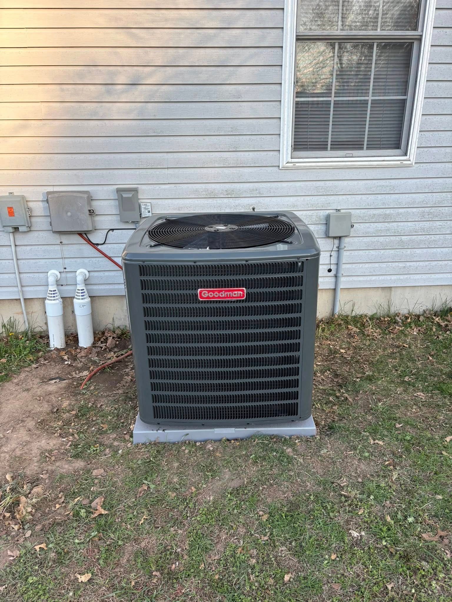 Gray Goodman air conditioner on a concrete base, beside a light-colored building with a window and electrical boxes.