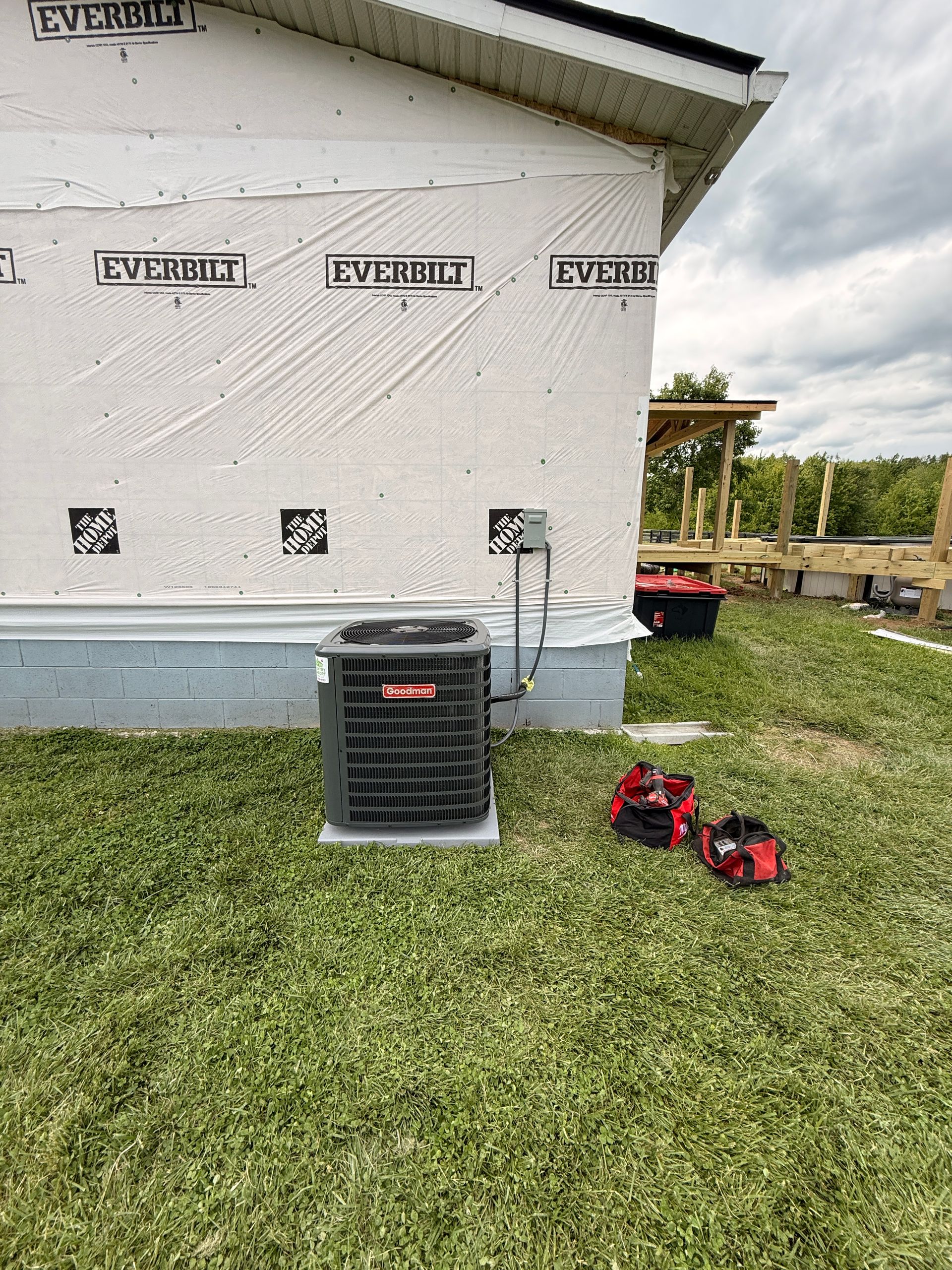Outdoor HVAC unit beside a house wall, with red tools on the grass and a ladder near the right side.