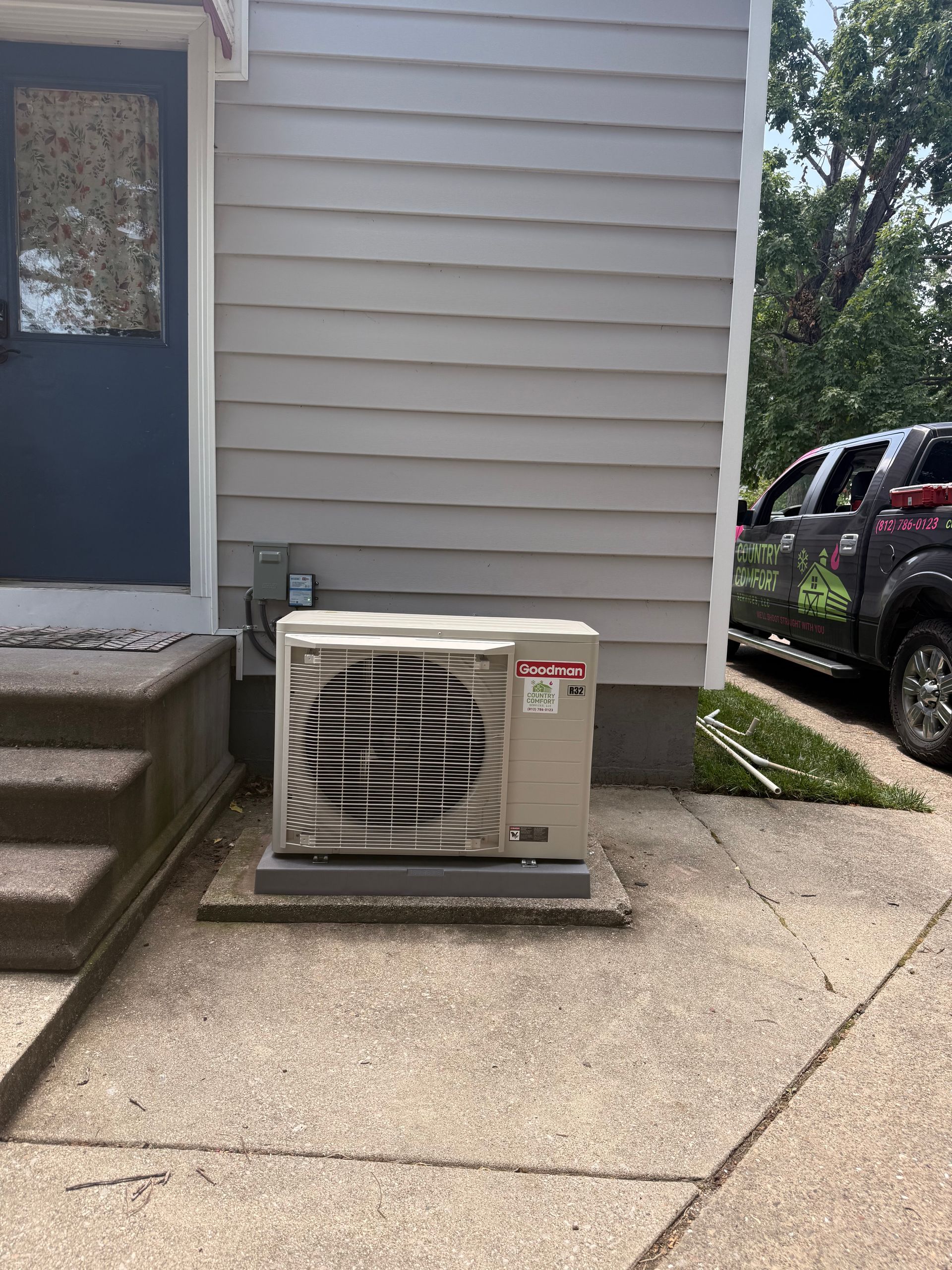 Outdoor air conditioner unit beside a beige house wall and concrete steps, with a car in the driveway.