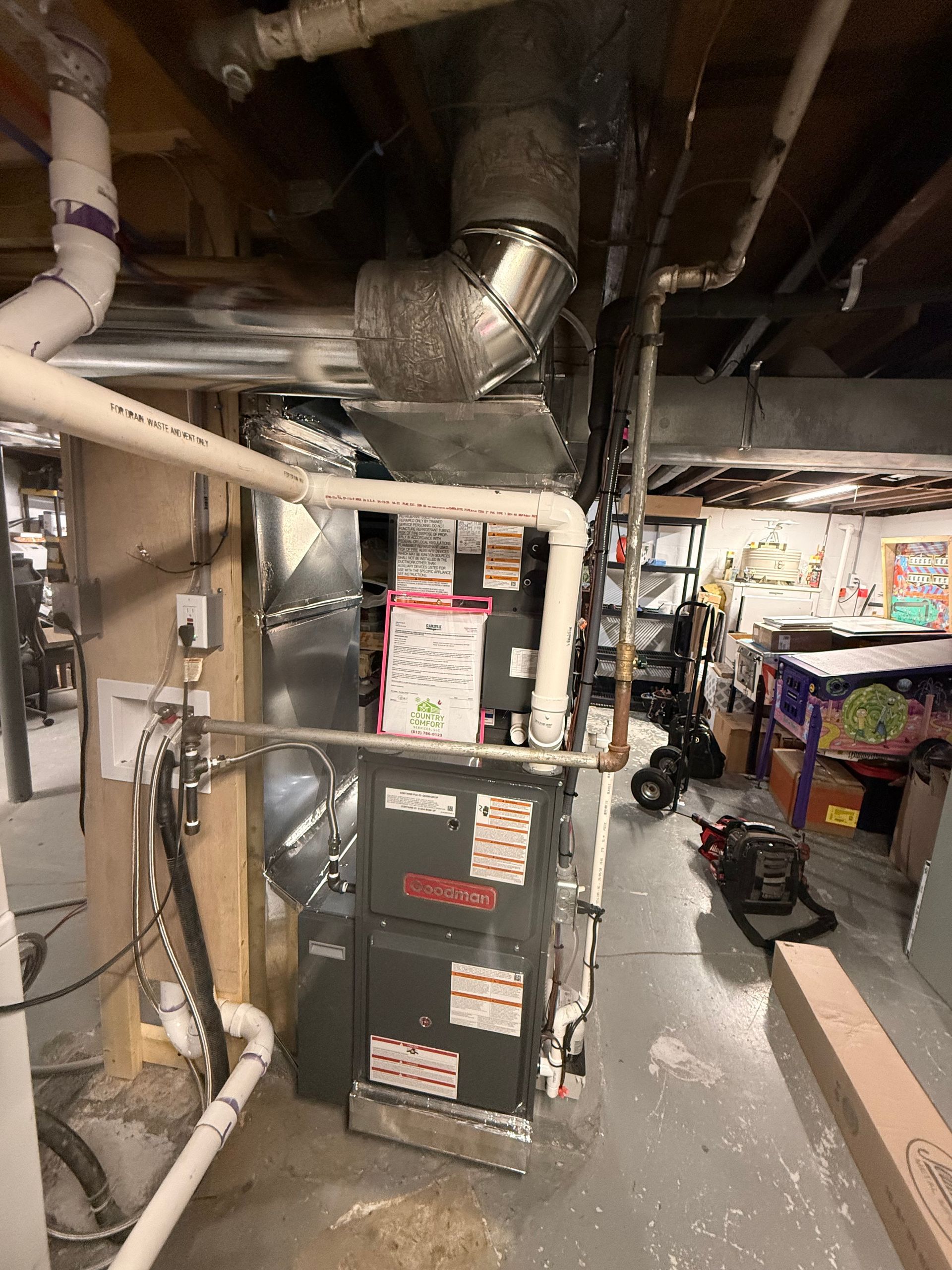 Basement furnace and HVAC equipment amid exposed pipes and ductwork, with storage items nearby