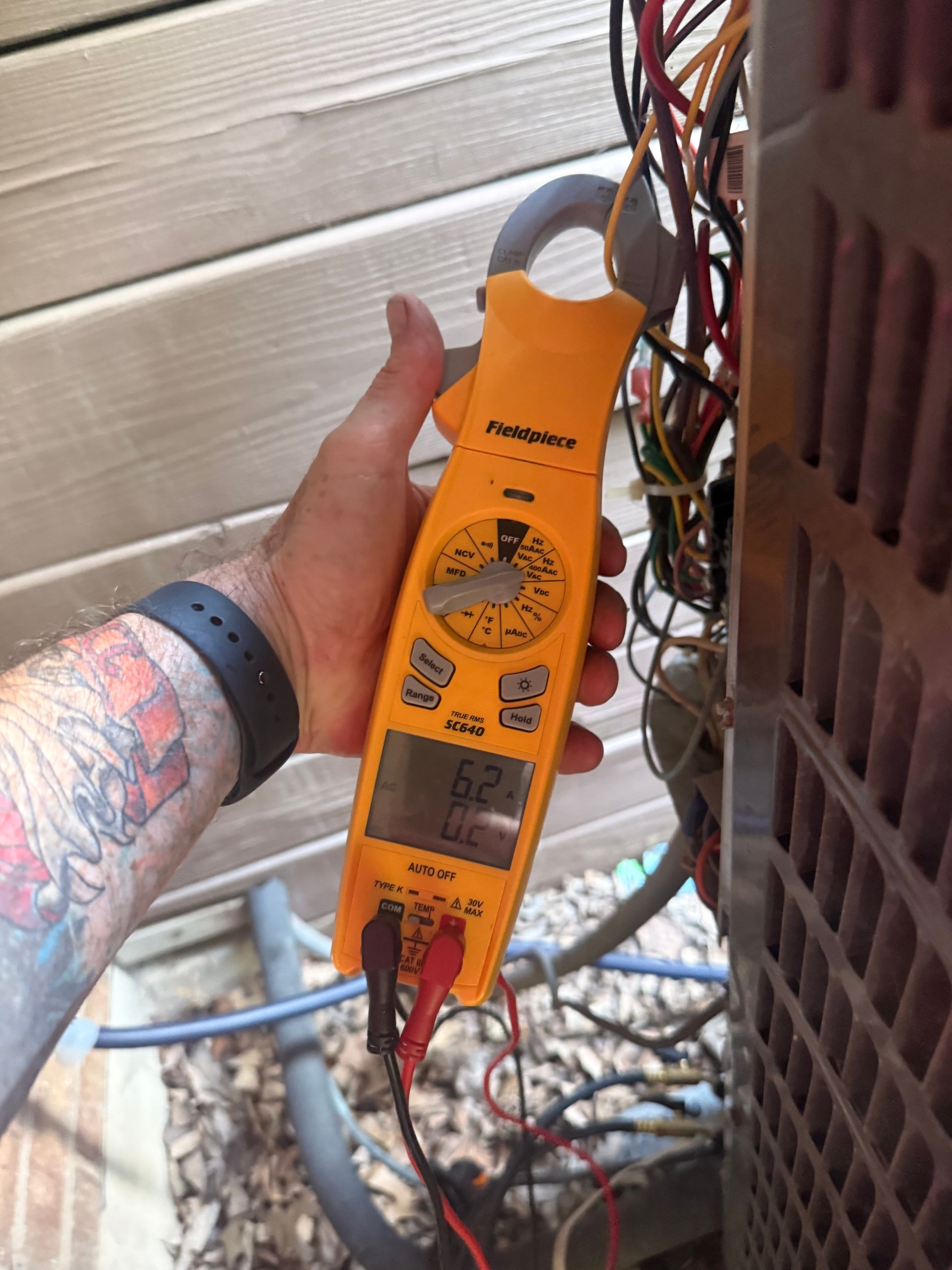 Hand holding an orange clamp meter near electrical wires beside an outdoor unit