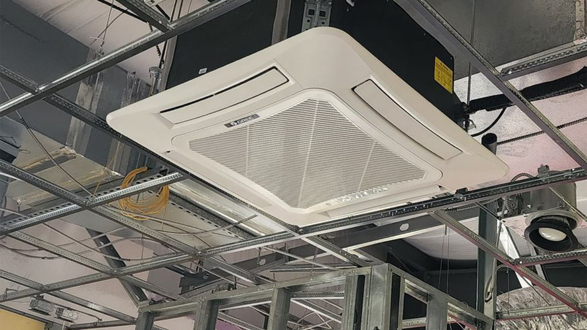 A white four-way ceiling cassette air conditioning unit mounted in an unfinished industrial ceiling.