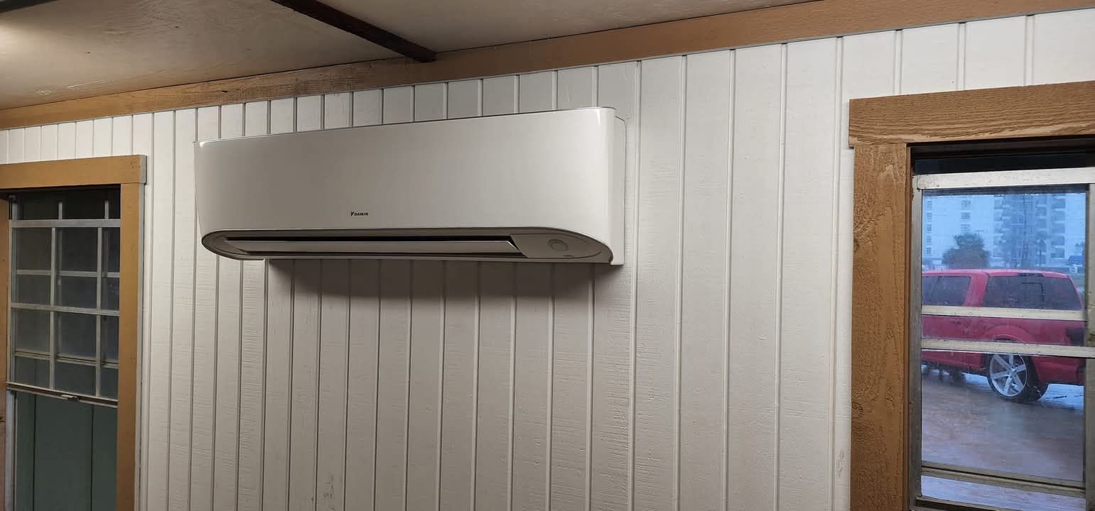 White wall-mounted air conditioner above wood-paneled wall between two windows.