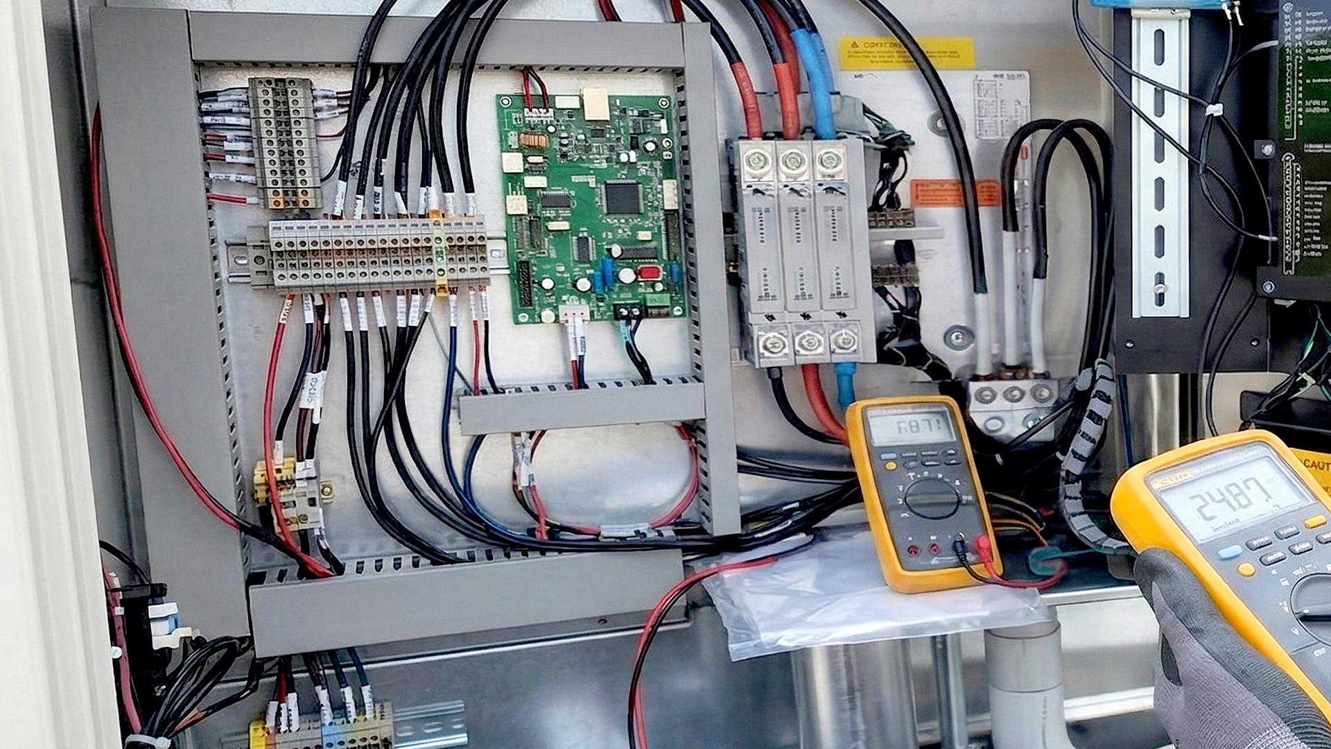 A person uses a yellow multimeter to test voltage on the wires inside an electrical control panel.