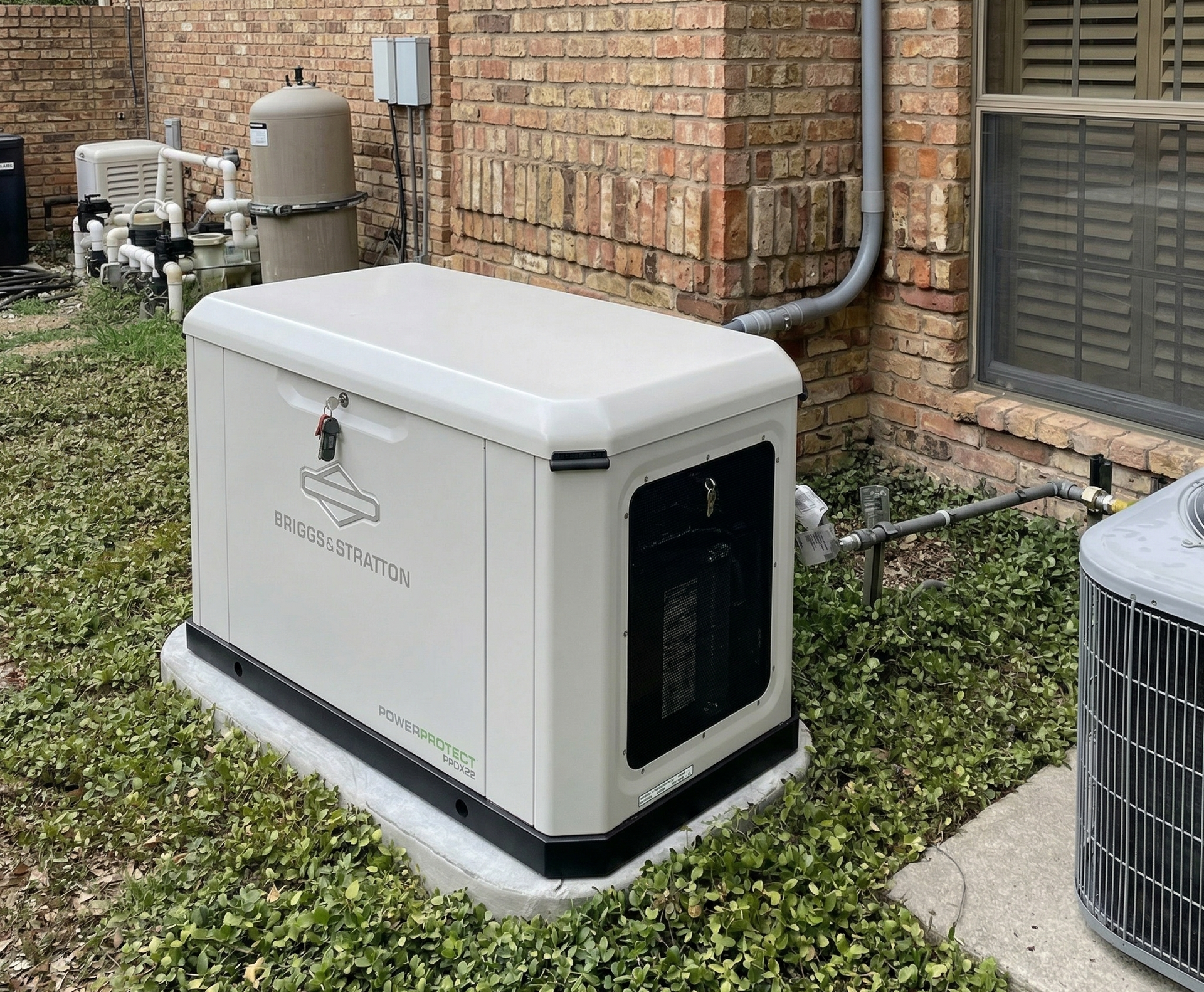 A beige residential standby generator sits on a concrete pad next to a brick house wall near air conditioning equipment.