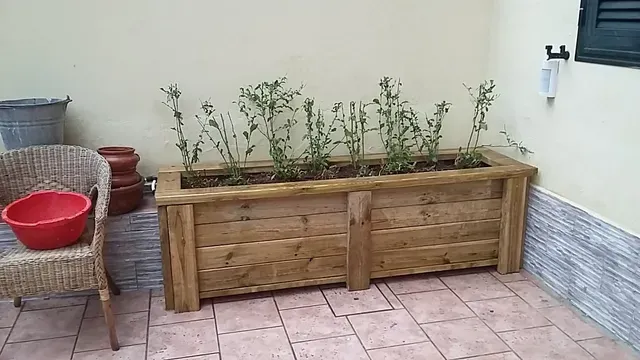 Fioriere in legno con poche piante su un patio piastrellato accanto a un muro, con un lavabo rosso su una sedia.