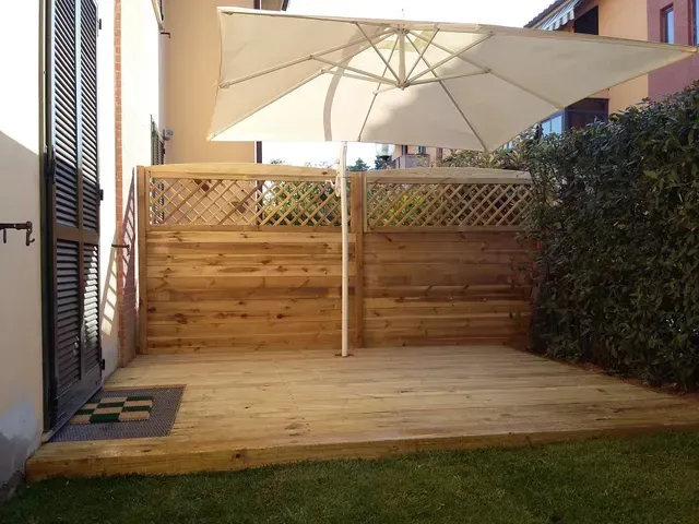 Terrazza in legno con un grande ombrellone bianco, recinzione a traliccio e siepe in un piccolo cortile sul retro.