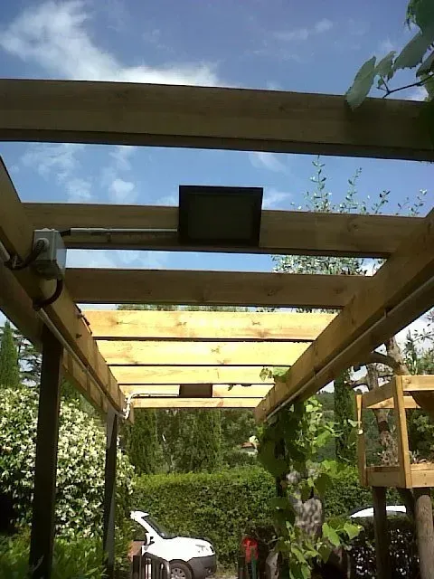 Un pergolato in legno incornicia un soleggiato patio in giardino con una sedia bianca, piante e il cielo azzurro sullo sfondo.