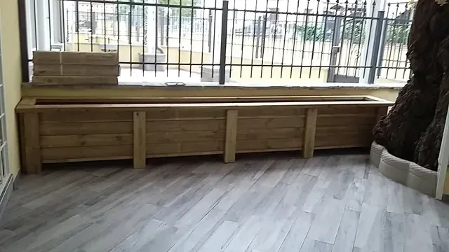 Panchina di legno addossata a una parete finestrata in una stanza luminosa con pavimento grigio.