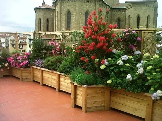 Fioriere in legno piene di fiori su una terrazza con un edificio in pietra sullo sfondo.
