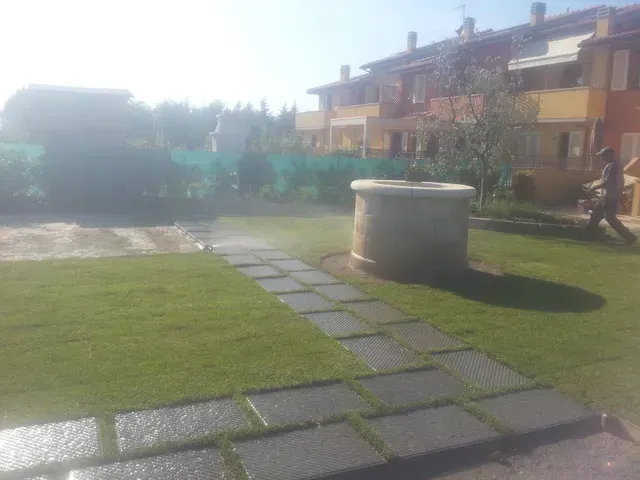 Cortile soleggiato con prato verde, sentiero in pietra e pozzo circolare in cemento vicino ai palazzi residenziali.