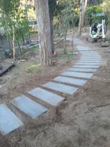 Sentiero sinuoso di pietre che attraversa un cortile alberato, con un piccolo escavatore sullo sfondo.