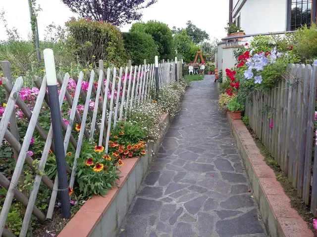 Stretto sentiero da giardino fiancheggiato da fiori, pali di recinzione bianchi e una staccionata di legno in una giornata nuvolosa.