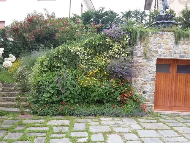 Casa in pietra con porta in legno, collina lussureggiante ricoperta di fiori e sentiero lastricato antistante.