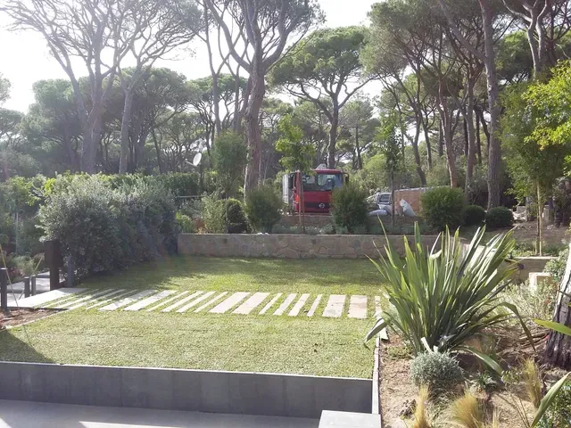 Giardino con prato, sentiero in pietra e alberi, con un veicolo rosso parzialmente visibile sullo sfondo.