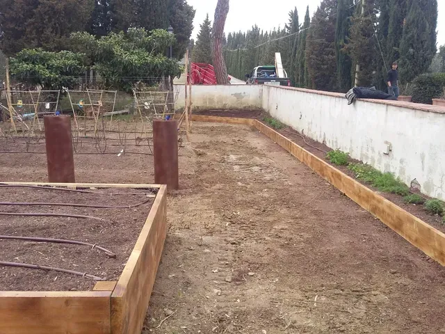 Un sentiero nel giardino accanto a aiuole rialzate, con un muro bianco, alberi e materiali da costruzione sullo sfondo.