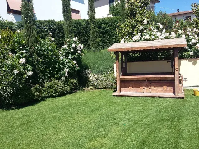 Giardino soleggiato con prato verde, arbusti in fiore bianchi e una zona salotto con pergolato in legno.