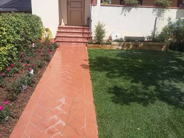 Vialetto d'accesso all'ingresso di una casa, con sentiero piastrellato di rosso, prato verde e cespugli lungo il lato.