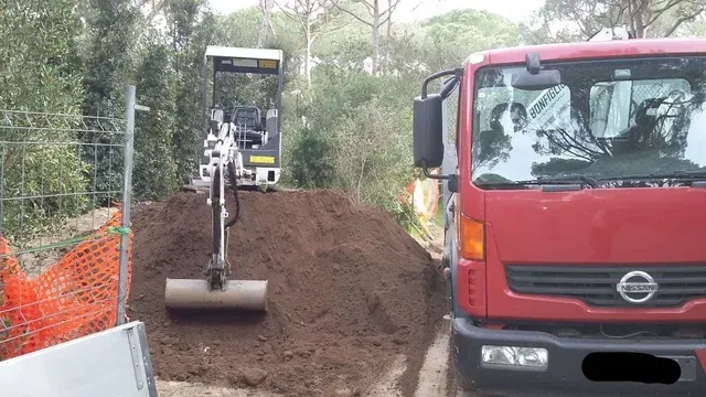 Un camion ribaltabile rosso e un escavatore stanno spargendo un cumulo di terra su una strada stretta.