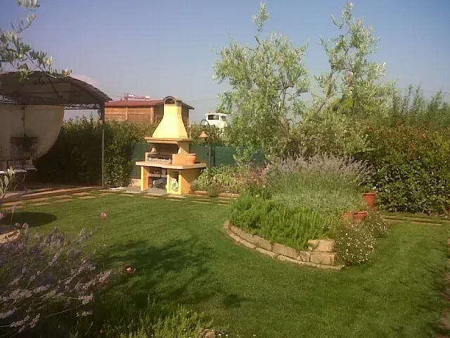 Giardino sul retro con prato verde, alberi, arbusti e un barbecue o caminetto da esterno di colore giallo.