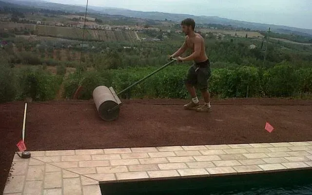 Un operaio fa rotolare un grosso cilindro su una terrazza sul tetto, con una verde vallata sullo sfondo.