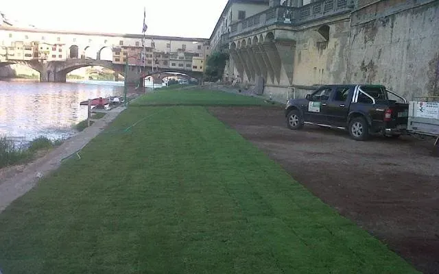 Sentiero lungo la riva del fiume, accanto a un terrapieno e a un ponte in pietra, con un SUV nero parcheggiato sulla destra.