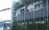 Esterno di un edificio industriale con rivestimento in metallo grigio, finestre e vegetazione rigogliosa sul davanti.