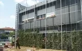 Un cantiere edile recintato con un grande edificio grigio e degli operai vicino a una fila di alberi.