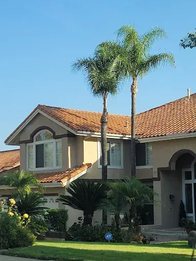 Two-story house with palm trees, brown tile roof, and beige exterior under a clear blue sky.
