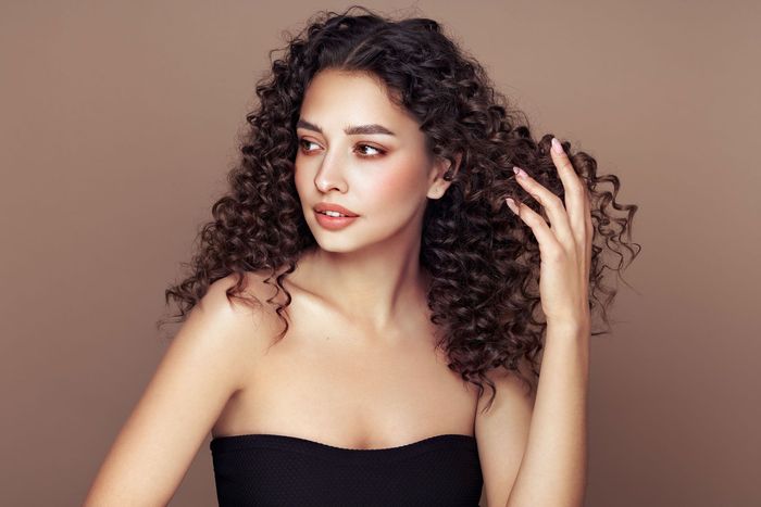 Woman with curly hair in a strapless black top, posing against a plain beige background.