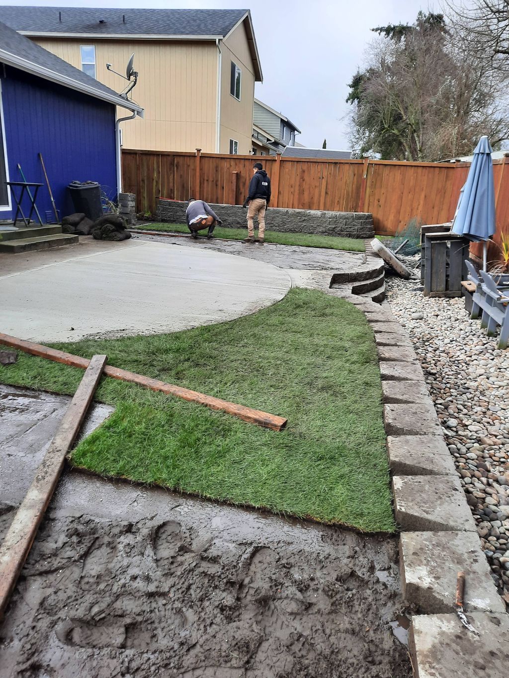 Backyard with muddy lawn, concrete patio, stone border, and two people near the wooden fence.