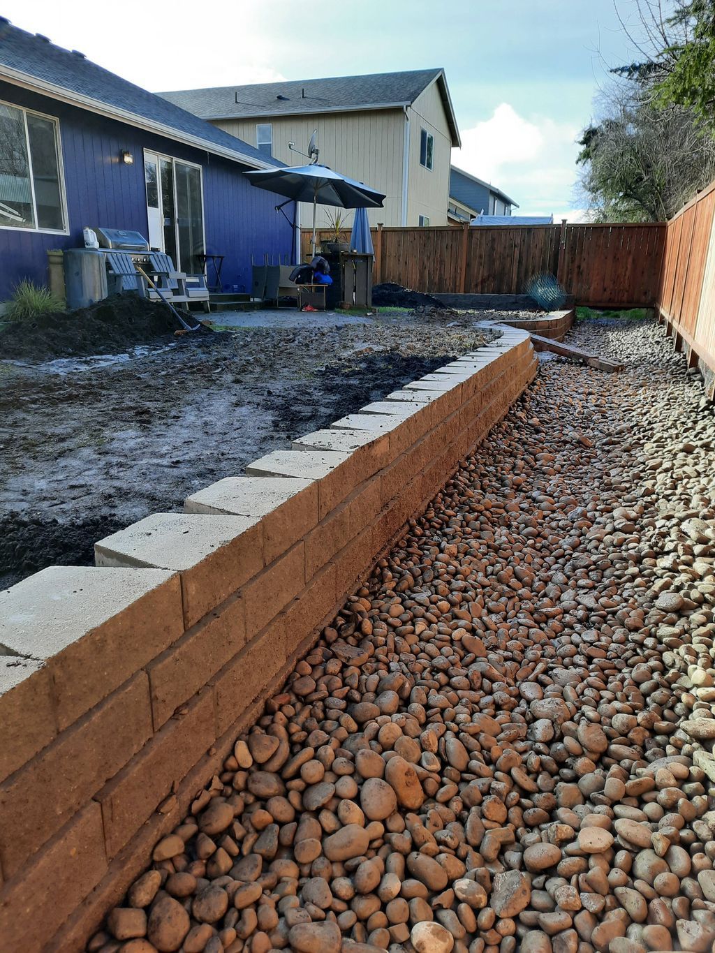 Backyard under construction with a retaining wall, gravel path, and blue house siding.