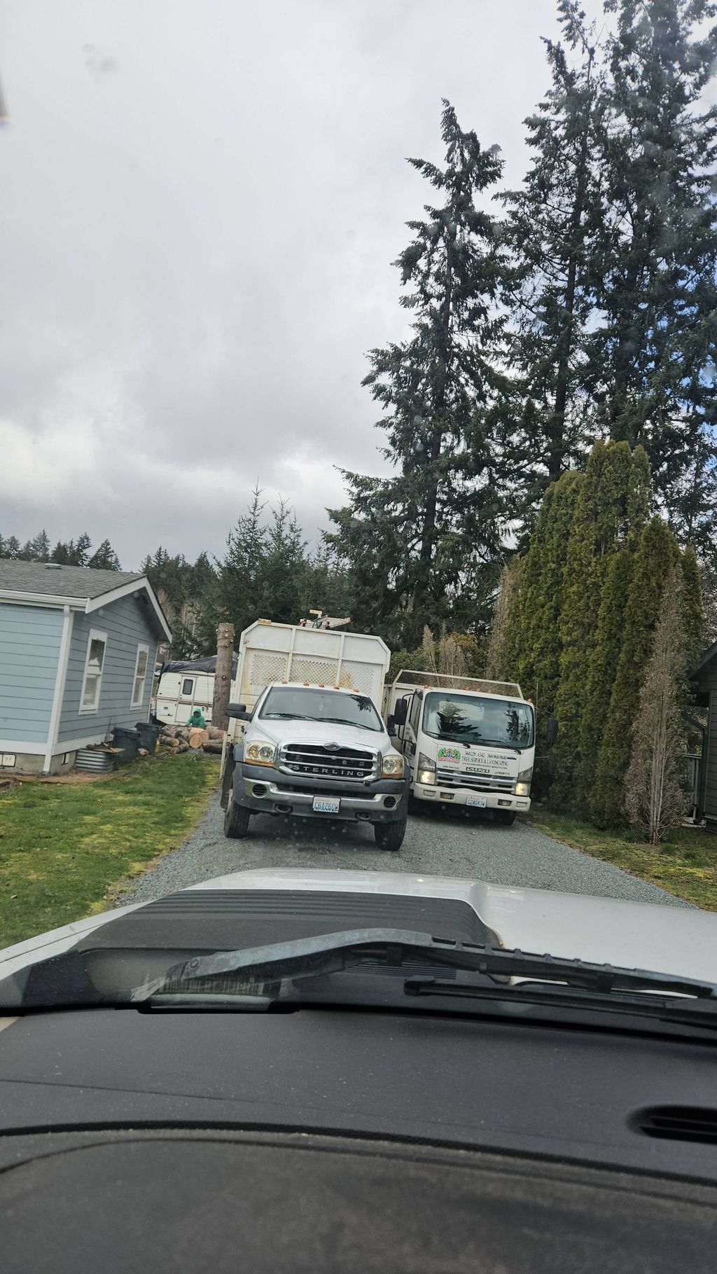 Rural driveway with two parked utility vans beside a house, viewed from inside a car under cloudy skies