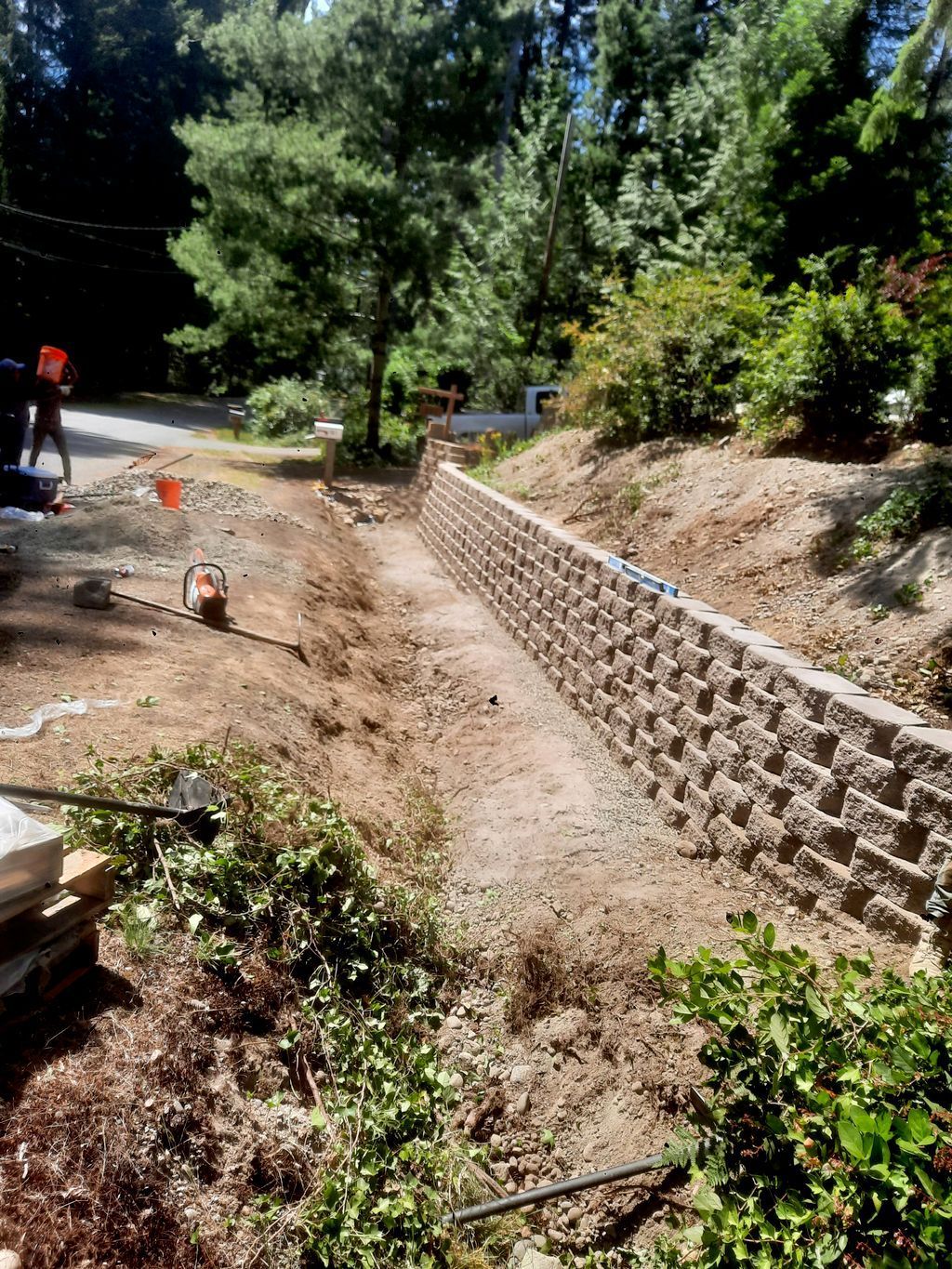Steep dirt drainage channel under construction beside a stone retaining wall in a wooded area.