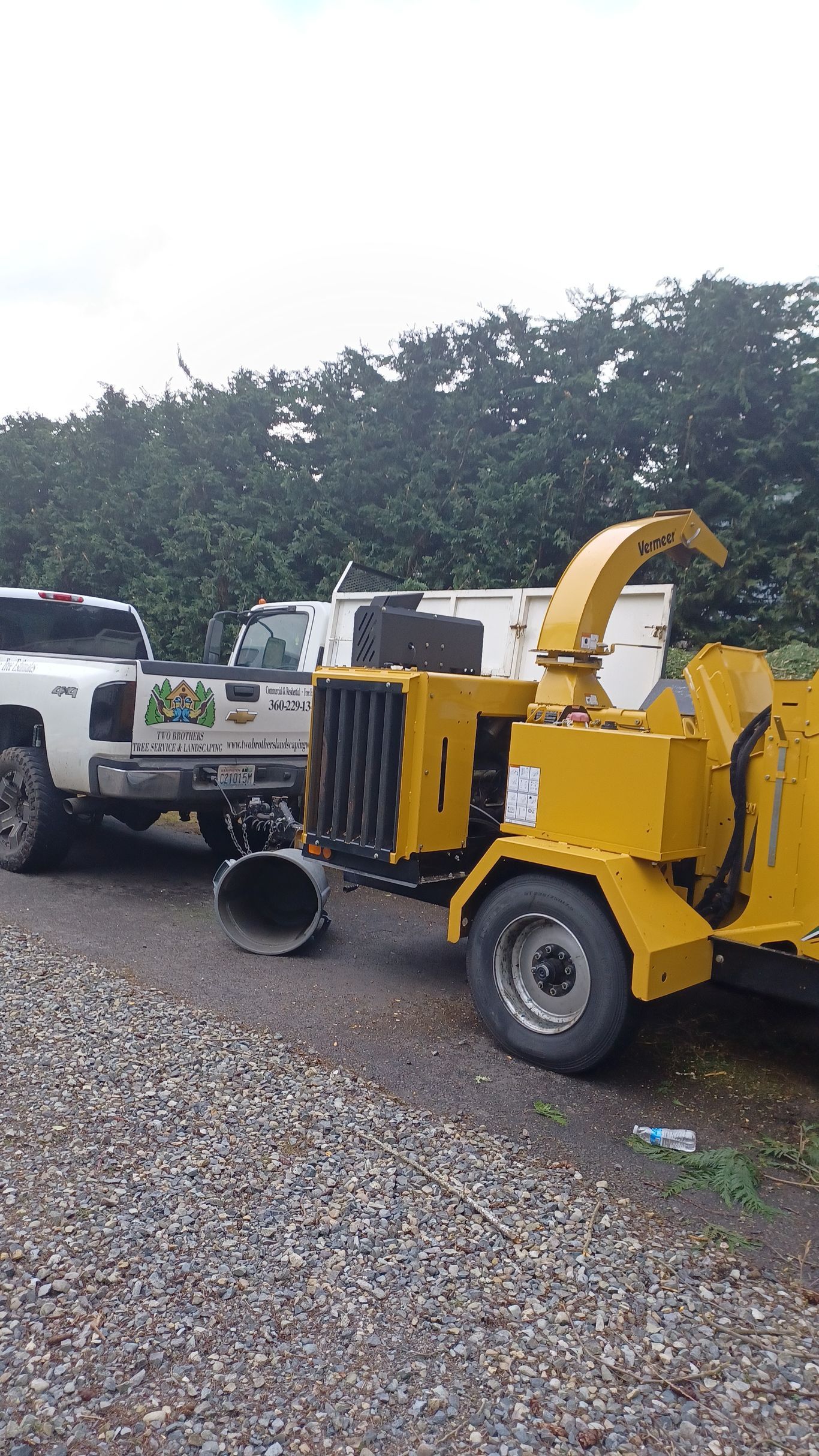 Yellow wood chipper parked on gravel beside a white utility truck, with trees in the background