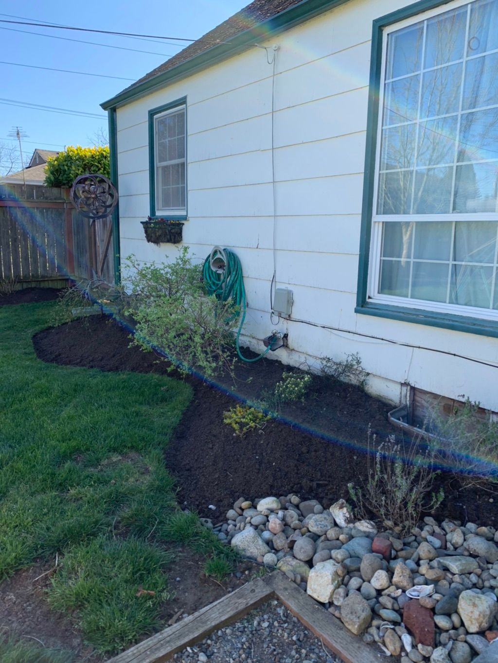 Side of a white house with a large window, garden hose, mulch bed, rocks, and a grassy yard.