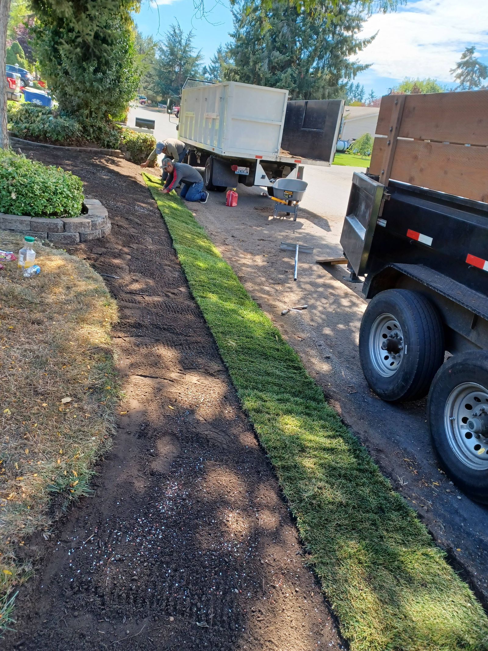 Landscaped yard with a small trailer and work truck beside a dirt driveway and tree-lined path