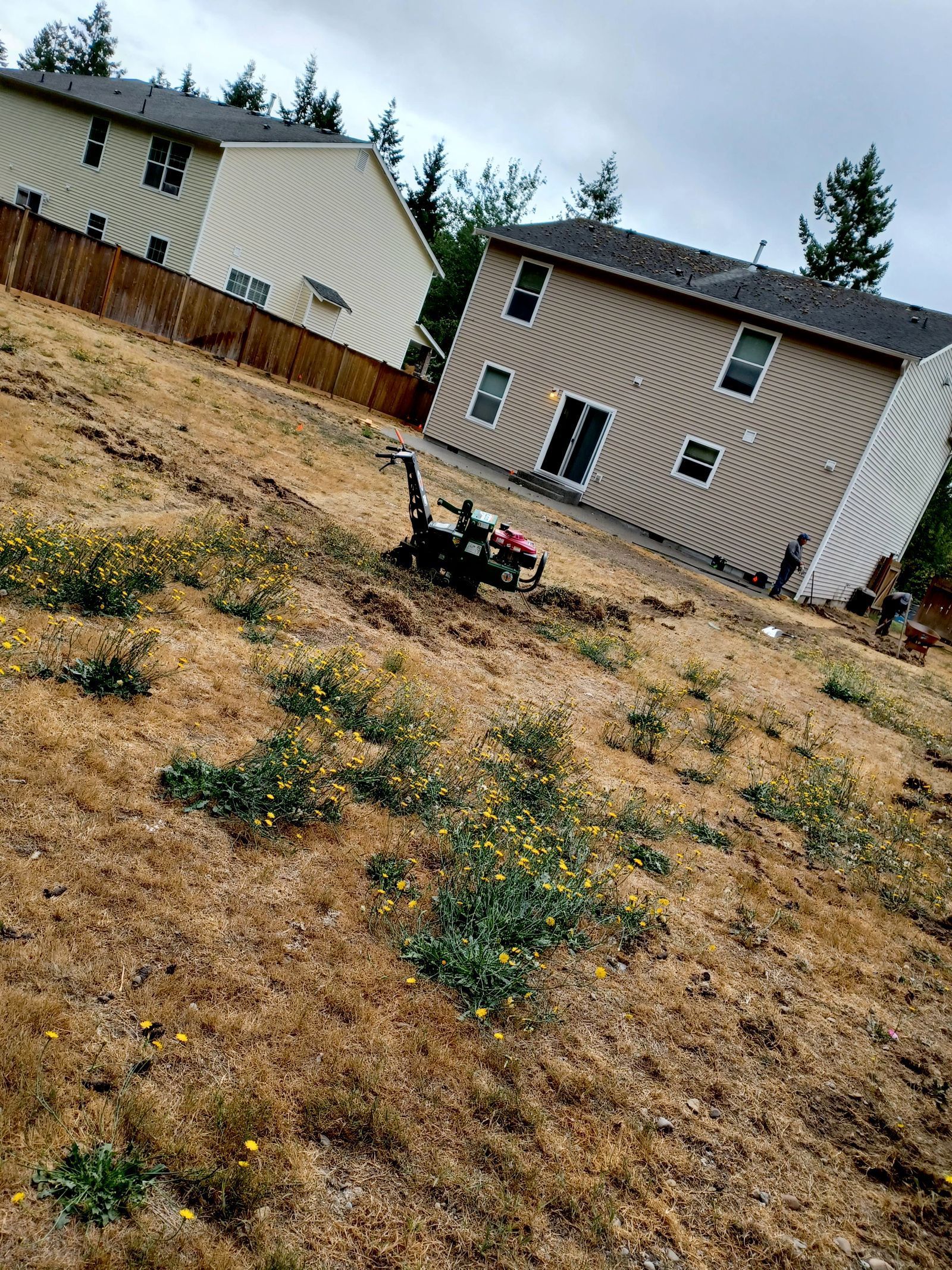 Two beige houses on a dry hillside, with a lawn mower in the foreground under cloudy skies