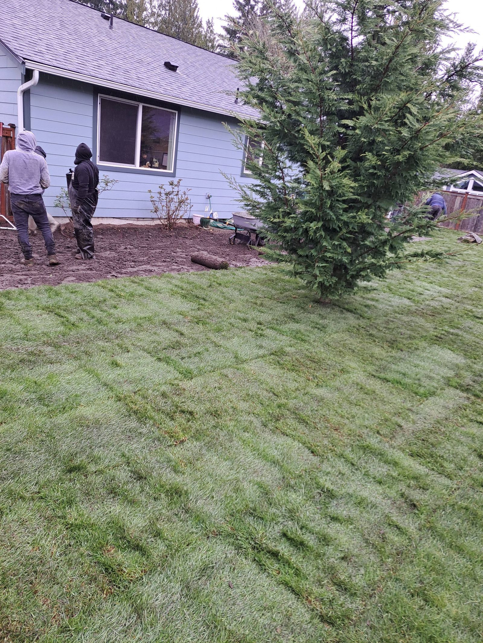 Two people stand by a house foundation beside a grassy yard and evergreen tree.