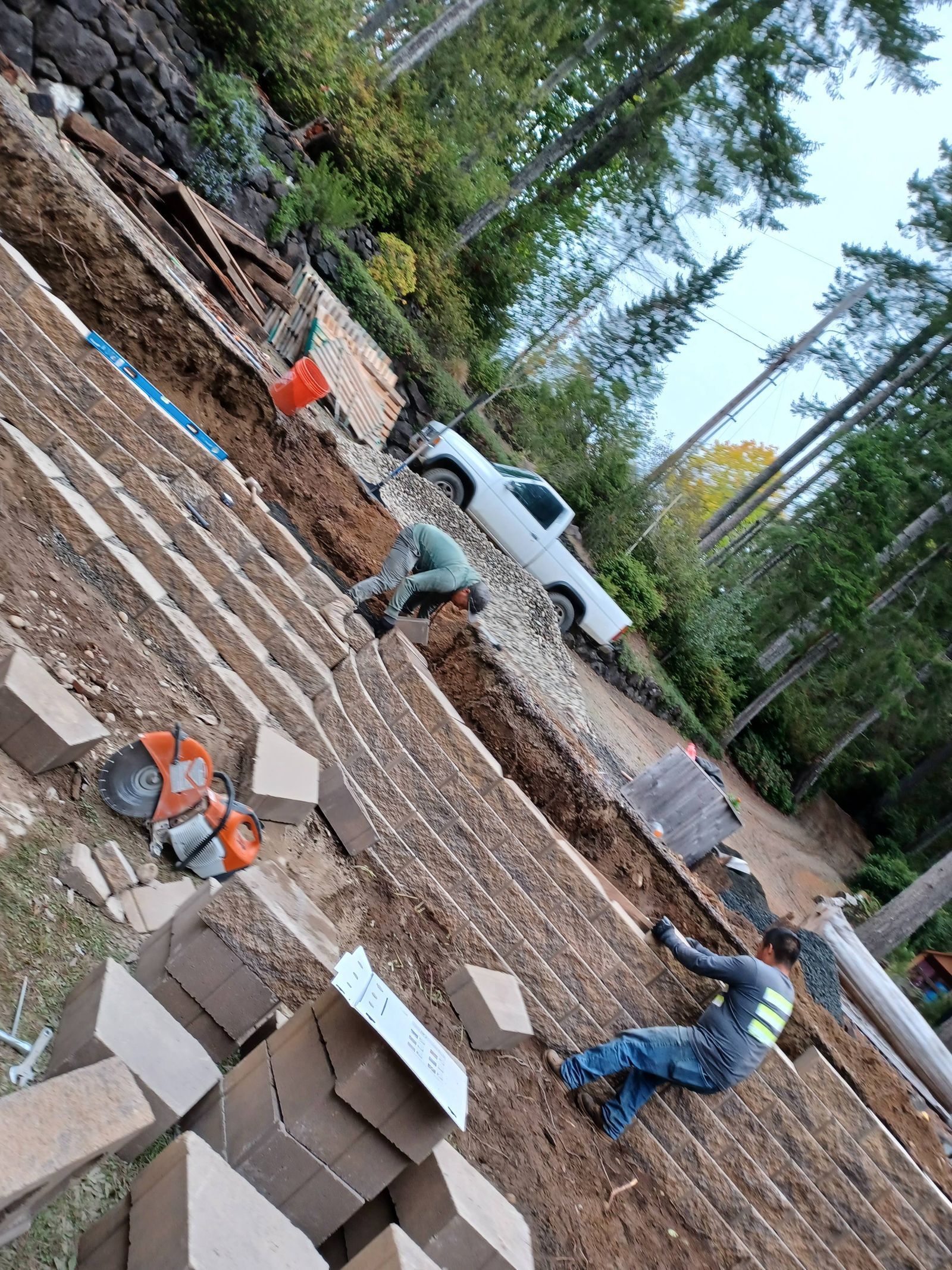Construction workers laying brick foundation beside a white pickup truck in a wooded outdoor site