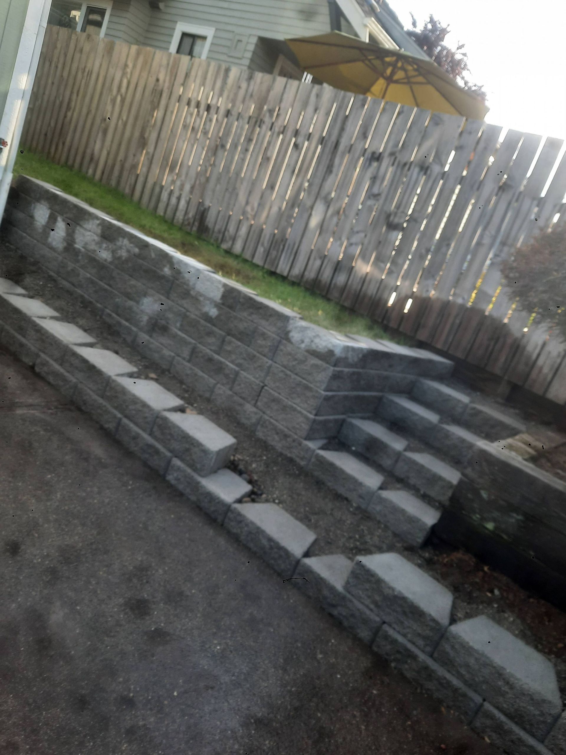 Tilted view of a stepped stone retaining wall beside a fence and driveway, with a patio umbrella above.