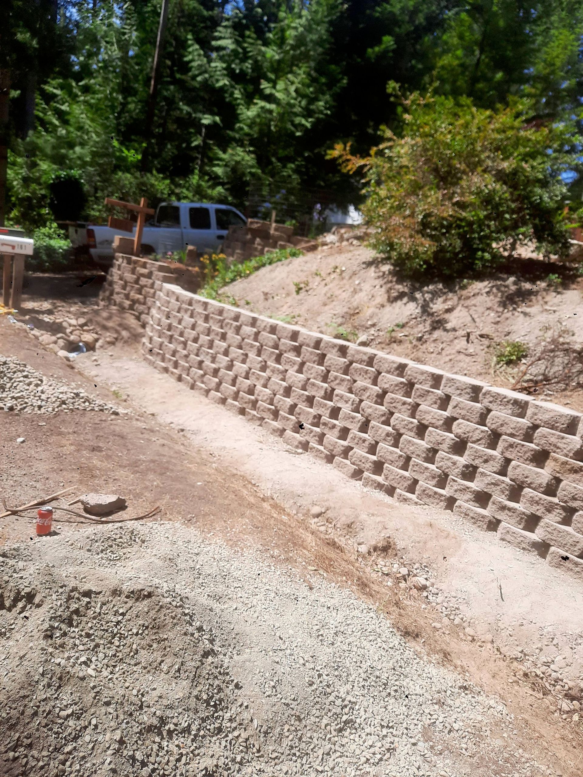 Gravel driveway beside a tan retaining wall on a wooded hillside, with a white truck in the background.
