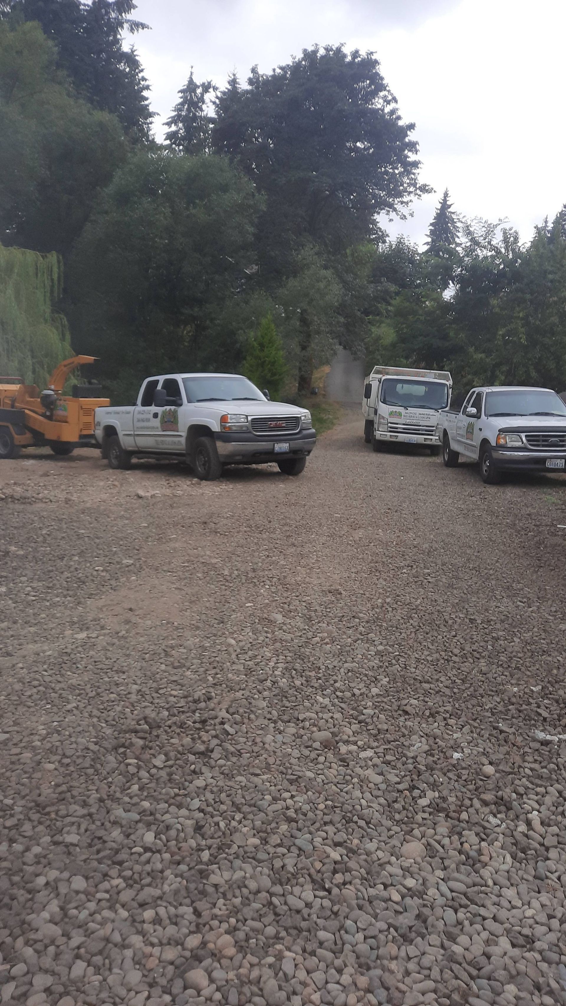 Gravel lot with parked pickup trucks and an excavator beside a wooded hillside