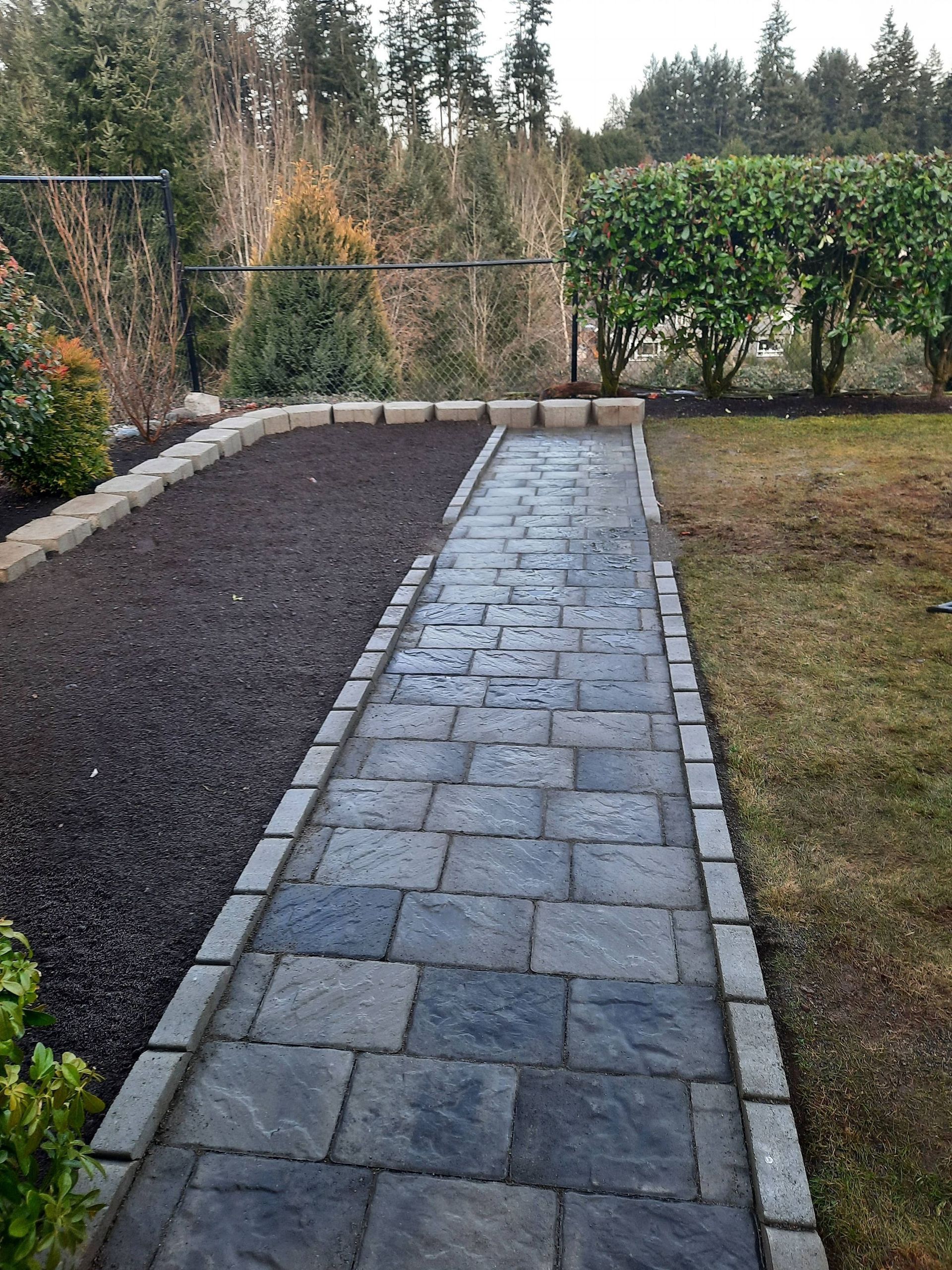 Stone walkway beside a dark gravel bed and lawn, leading to a wooded backyard.