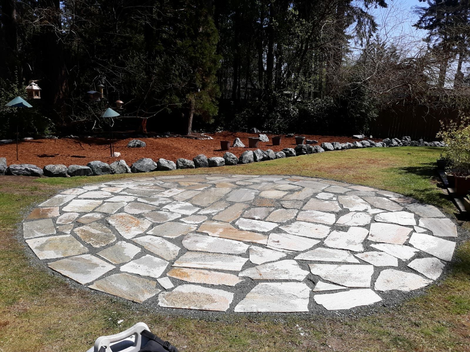 Circular stone patio in a grassy yard with trees and a flower bed in the background