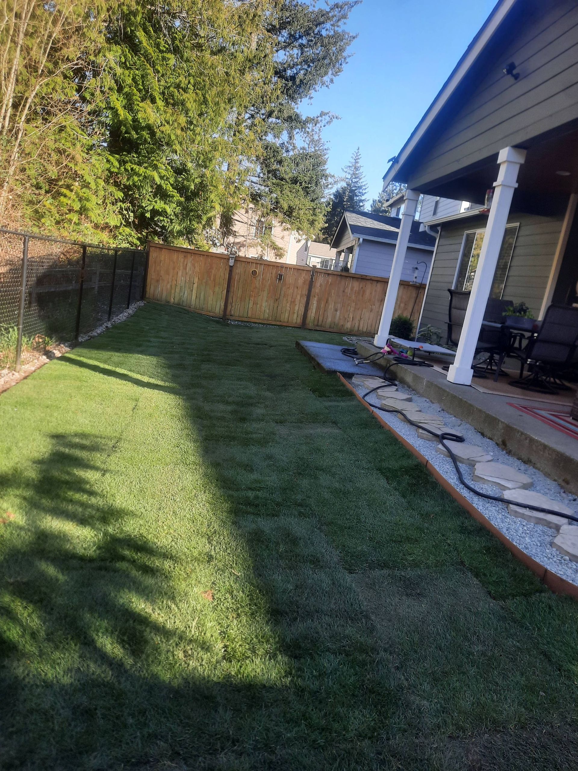 Backyard lawn beside a house porch, with a wooden fence and trees in morning sunlight